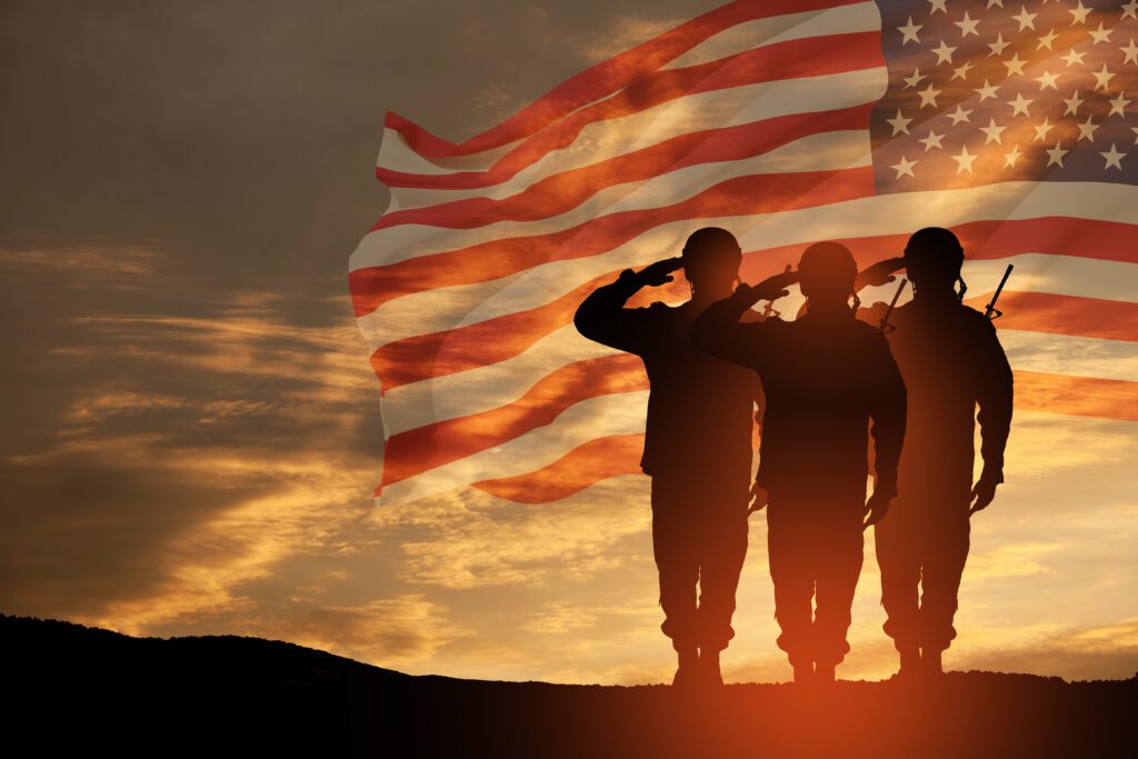 The silhouette of 3 soldiers saluting at sunset with an American flag in the background.